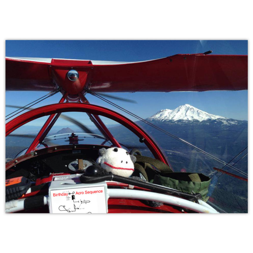 a sock monkey rides in the front seat of a red Pitts bi-plane with snow-capped mountains in the background